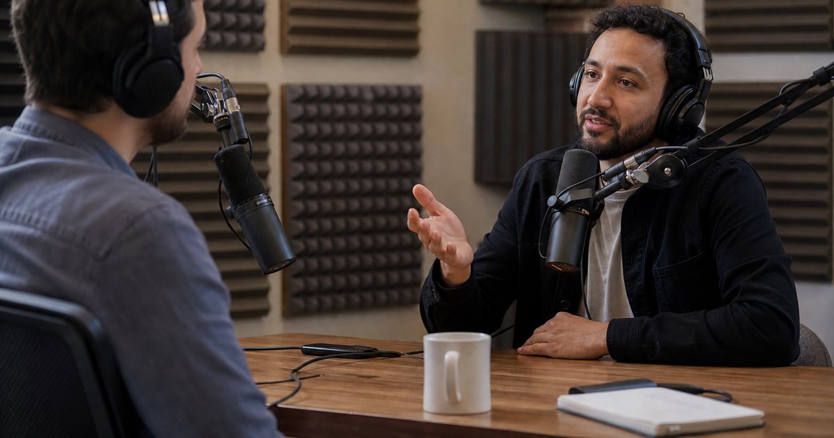 Two podcasters seated at a small studio desk with microphones on boom arms, headphones on, one gesturing mid-conversation, warm acoustic foam panels on the wall behind them