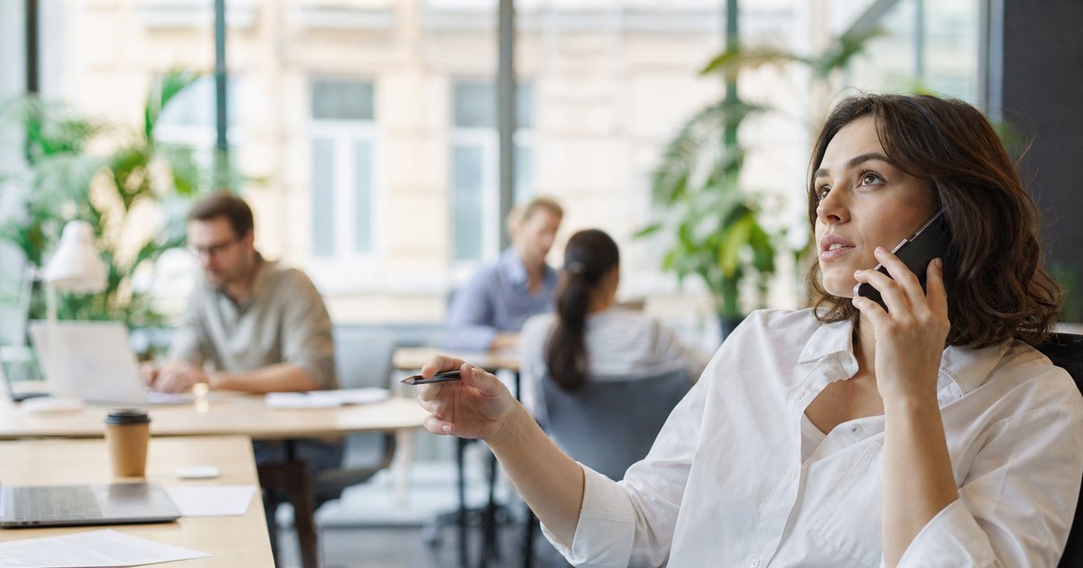 A newsletter writer in a bright coworking space on a phone call, leaning back in a chair with a pen in hand, colleagues blurred in the background, large windows and plants